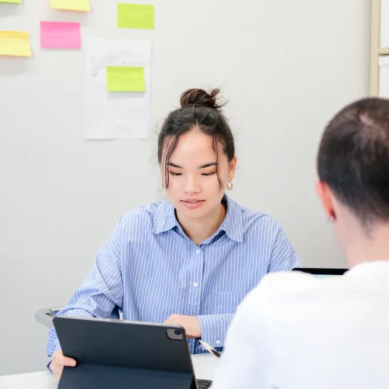 Junge Frau sitzt vor dem Laptop. An der Wand kleben Post-it in verschiedenen Farben. Der Rücken eines Mannes 