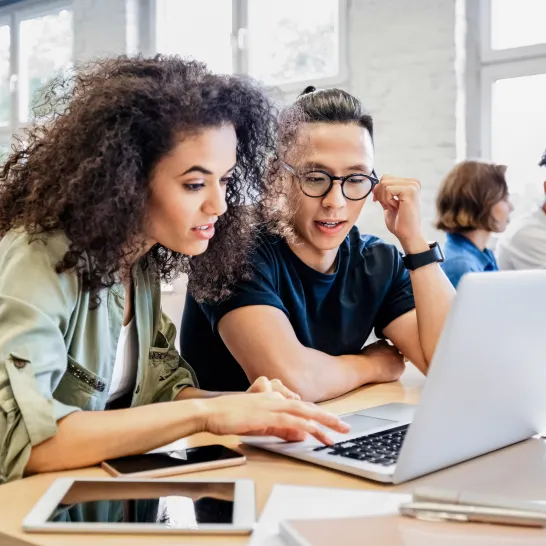 Eine junge Frau mit Locken und ein junger Mann mit Brille schauen in einen Laptop