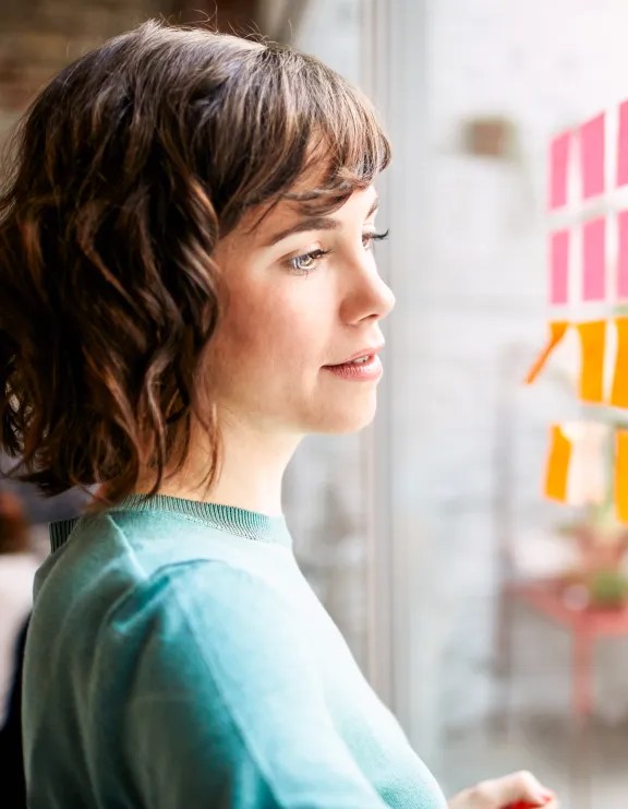 Frau steht vor einer durchsihtigen Wand, die mit Post-it beklebt ist und sie schreibt auf die Wand
